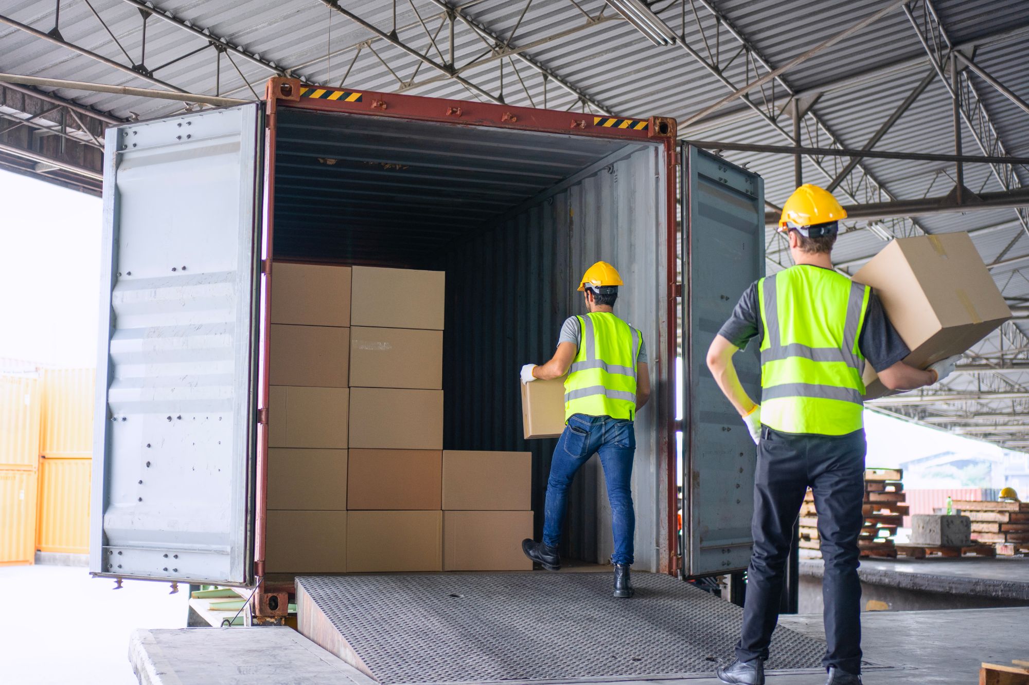 Dos operarios con casco y chaleco cargando cajas en un contenedor abierto sobre la rampa del muelle