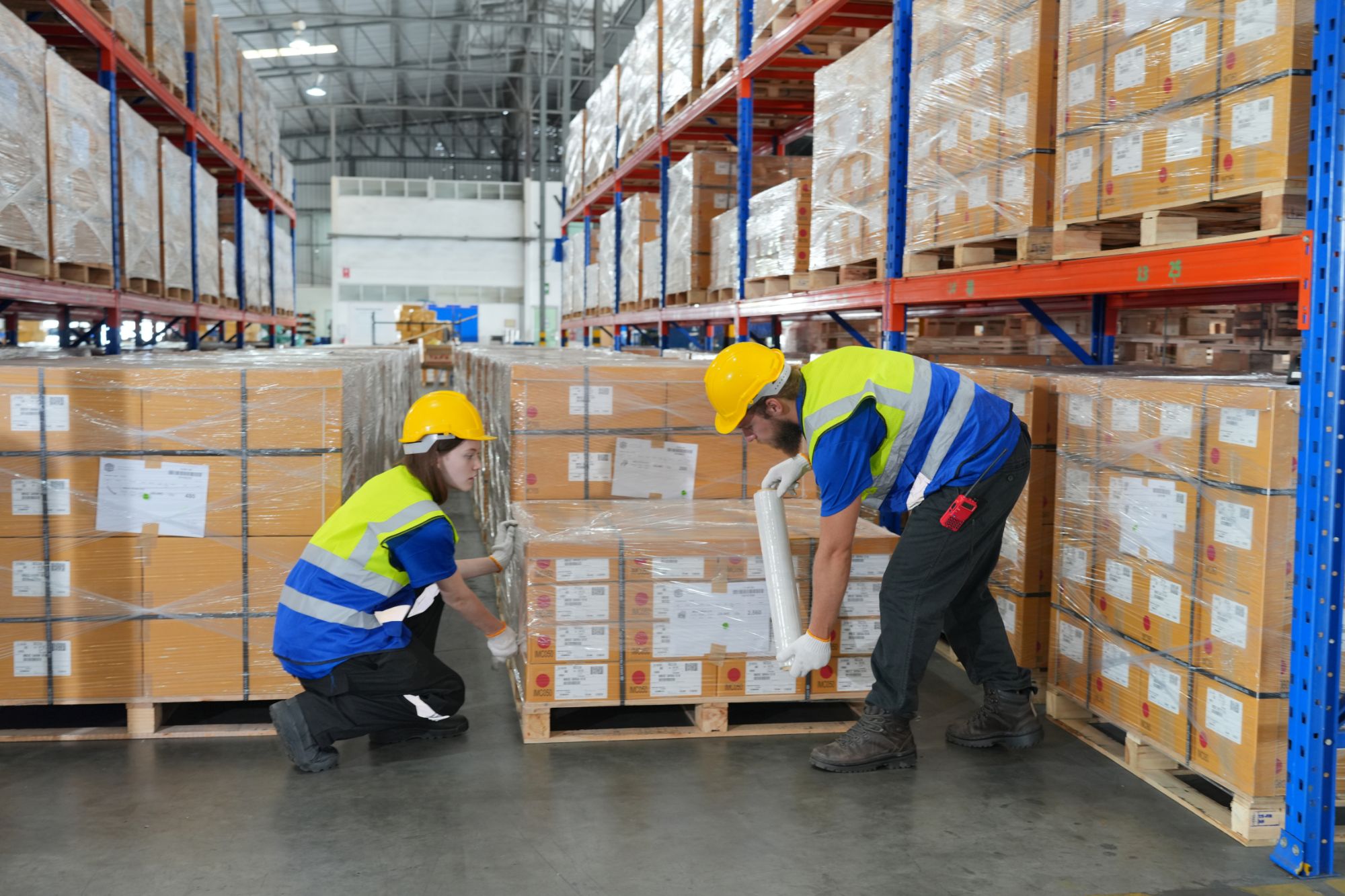 Dos operarios con casco y chaleco enfilmando con film estirable un pallet de cajas en un pasillo del depósito
